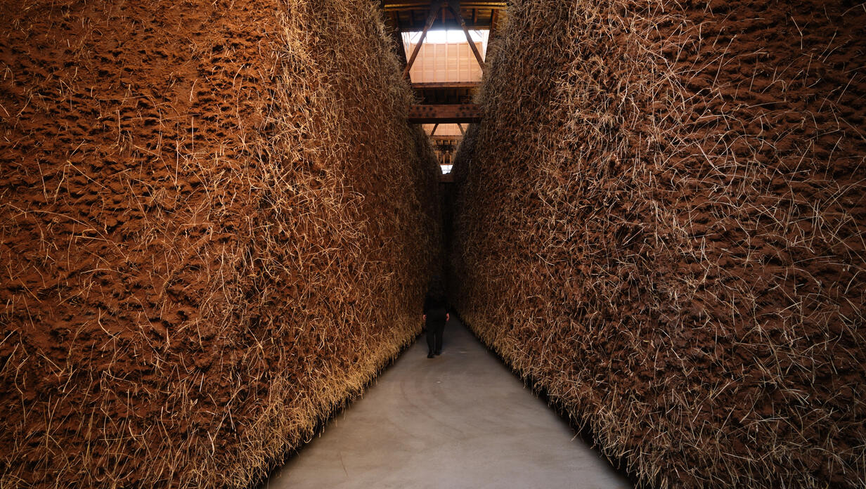 A narrow pathway flanked by tall walls made of straw or hay bales. The ground is a smooth surface, and the space is dimly lit, creating a sense of isolation.