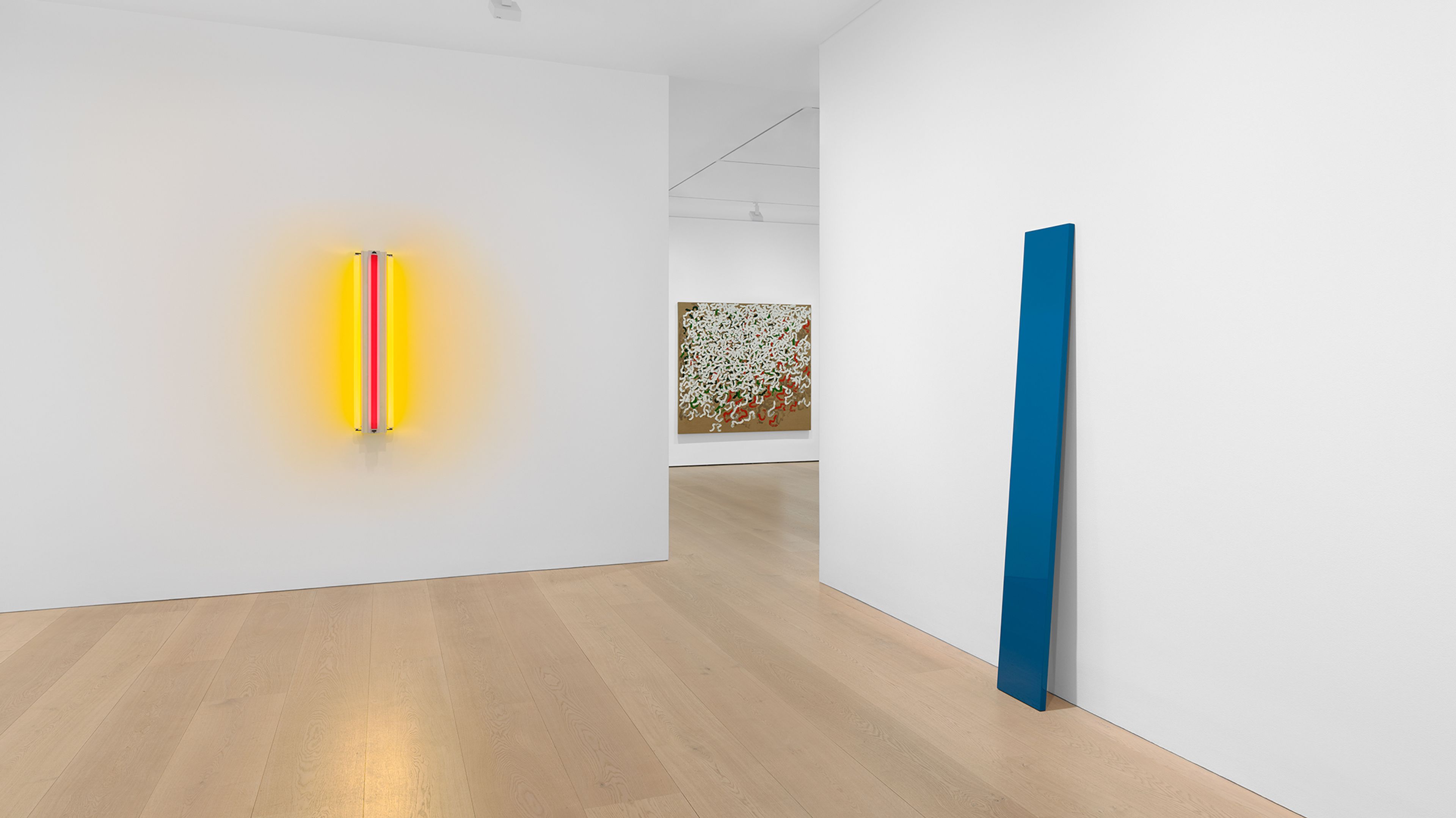 An interior gallery space with white walls and wooden flooring. On the left, a vertical neon light installation featuring yellow and red hues. In the background, a piece of artwork with colorful abstract patterns can be seen. To the right, a tall blue vertical sculpture stands against the wall.