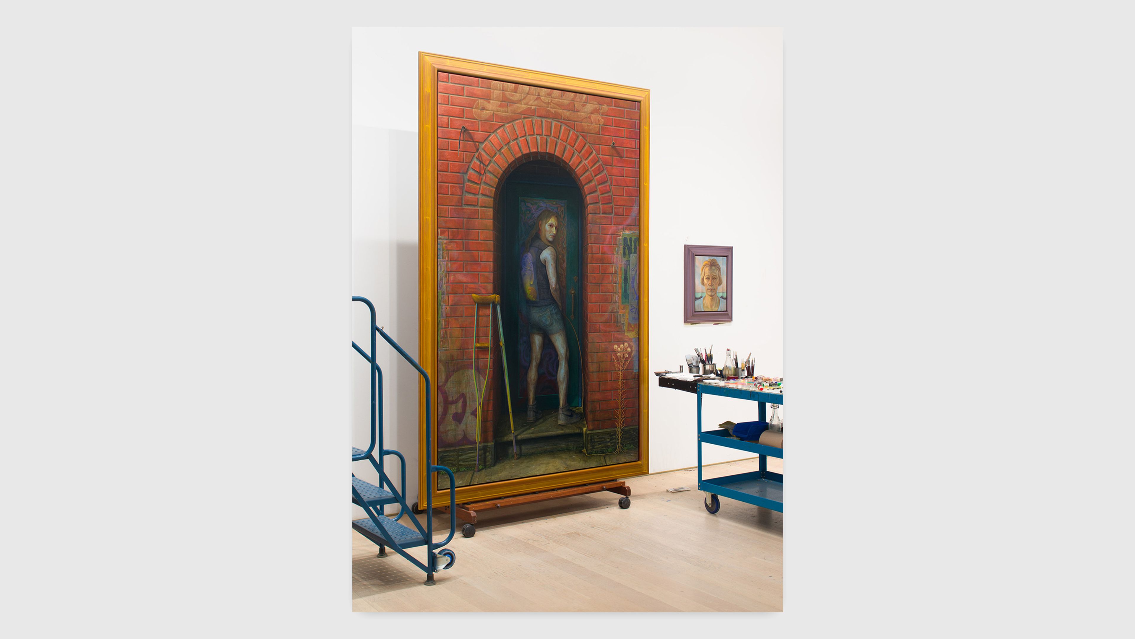A large painting of a figure standing in an archway, set against a brick wall, displayed in an art gallery with various tools and supplies nearby.