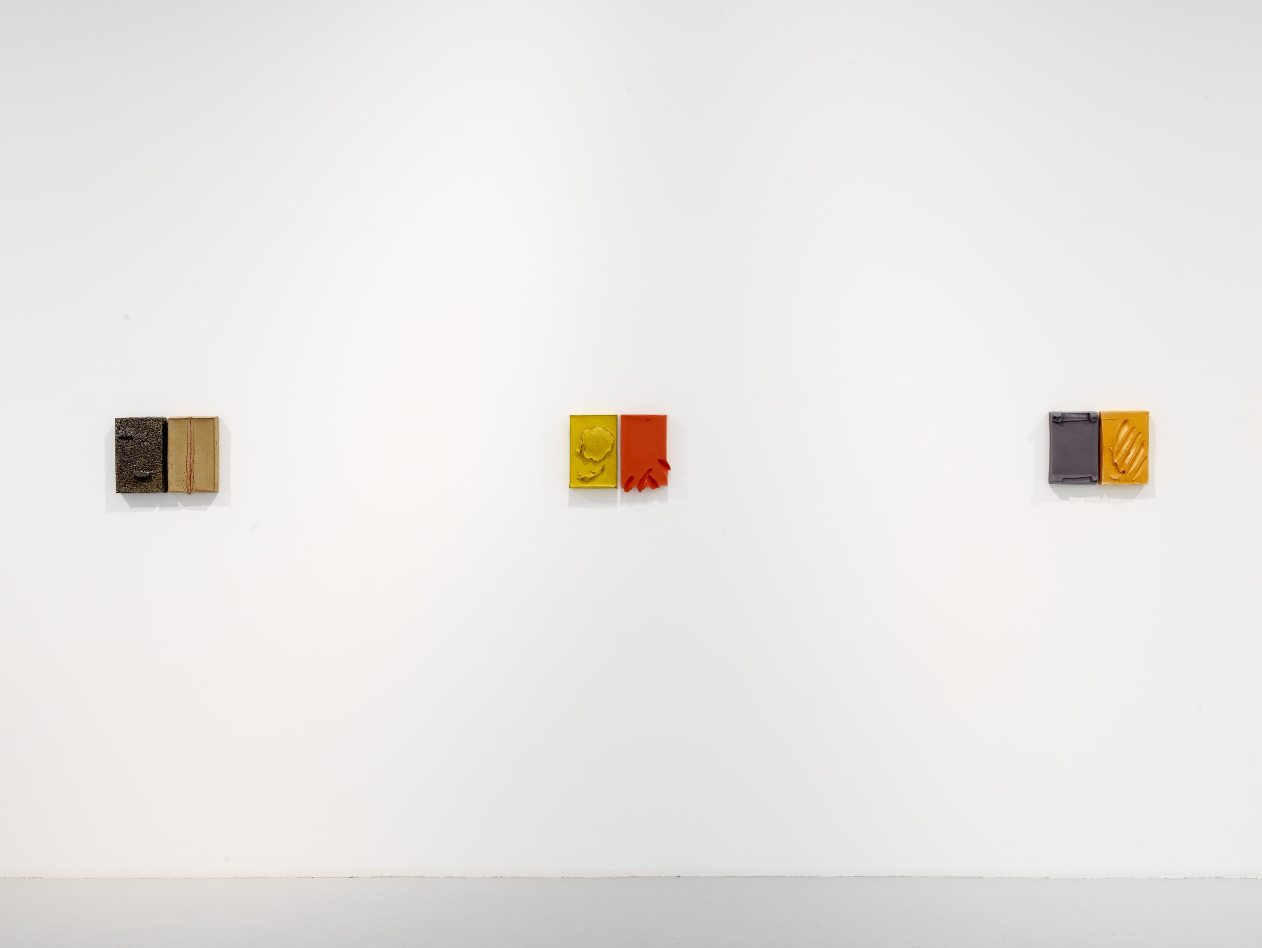 Three small artworks displayed on a white wall, arranged in a horizontal line. The artwork colors include brown, yellow, red, and gray.