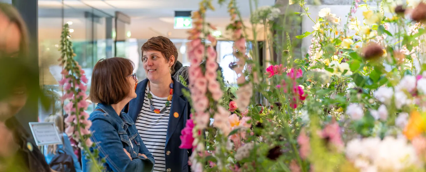 Two women talking amidst a display of colorful flowers in a bright, airy space.