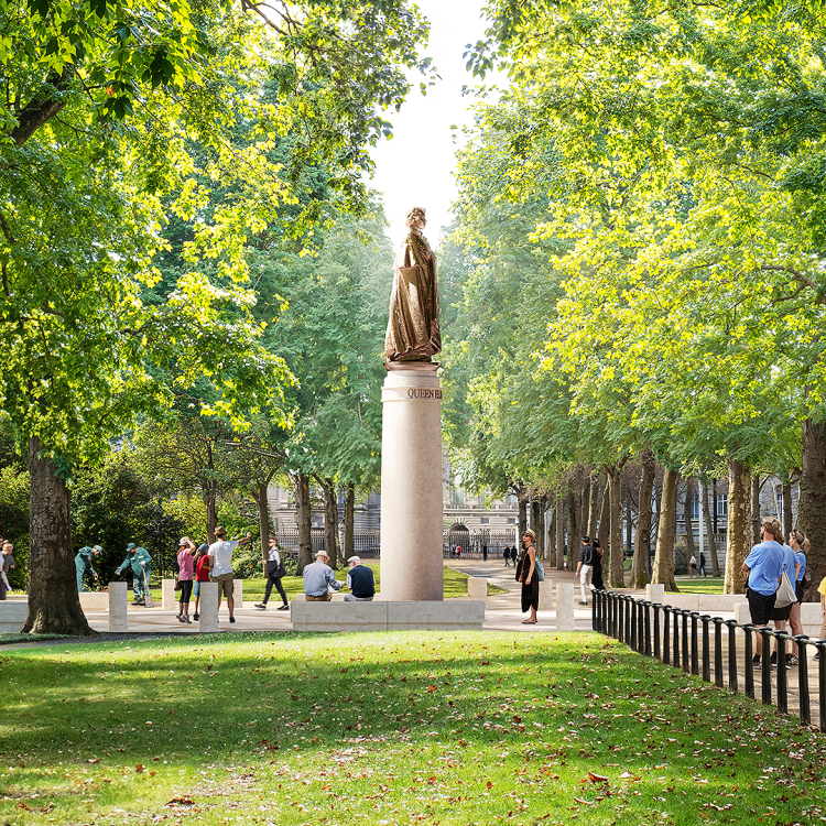 A statue of a figure wearing robes stands atop a tall pedestal in a park, surrounded by trees and people walking along paths.