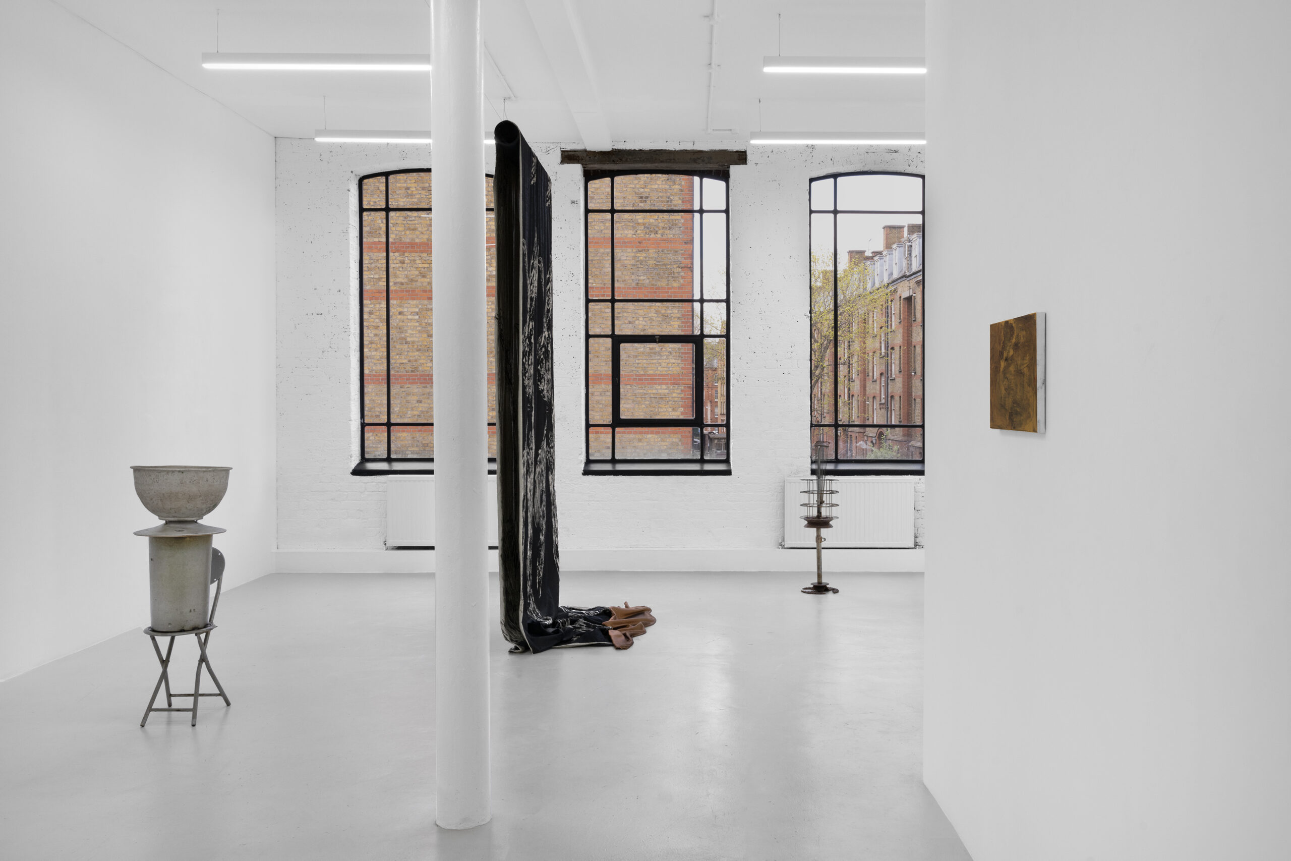A modern art gallery featuring white walls, large windows, and minimalist installations including a sculpture, a draped fabric, and a small artwork on the wall.
