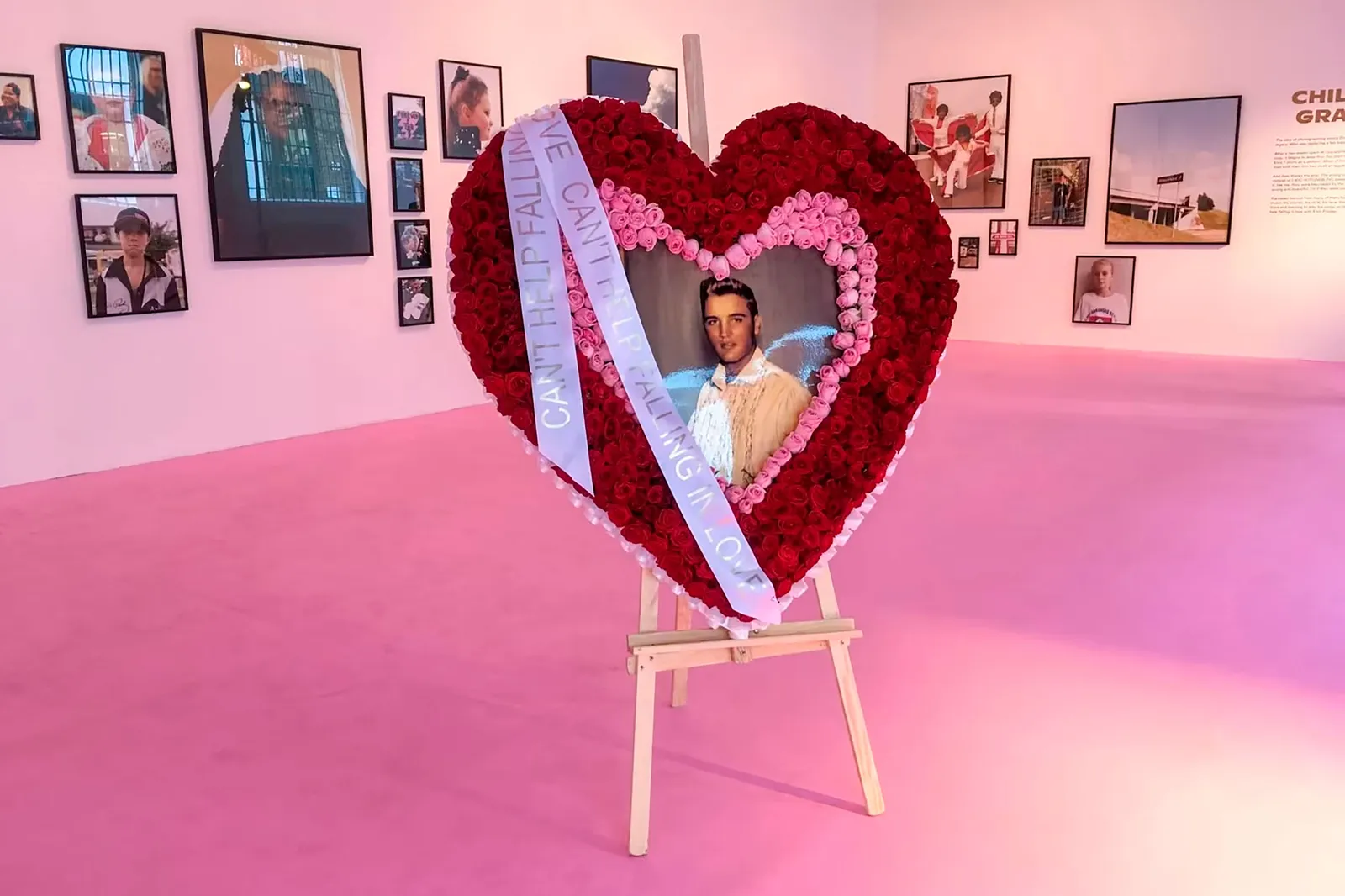 A heart-shaped floral tribute with a central photograph and ribbons, surrounded by framed pictures on the walls of an art gallery featuring pink flooring.