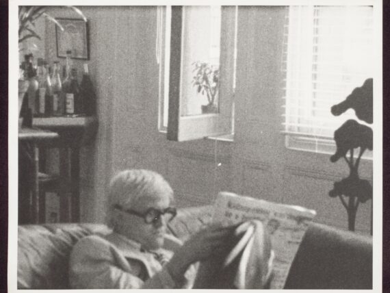 A black and white photograph of a person with gray hair sitting on a couch, reading a newspaper in a room with light coming through the window. There are bottles on a shelf in the background.