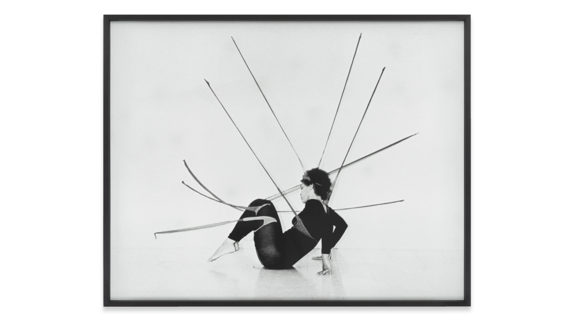A black and white photograph of a person sitting on the floor, with several long sticks protruding from their body, creating a striking visual effect. The background is plain, enhancing the focus on the figure and the sticks.