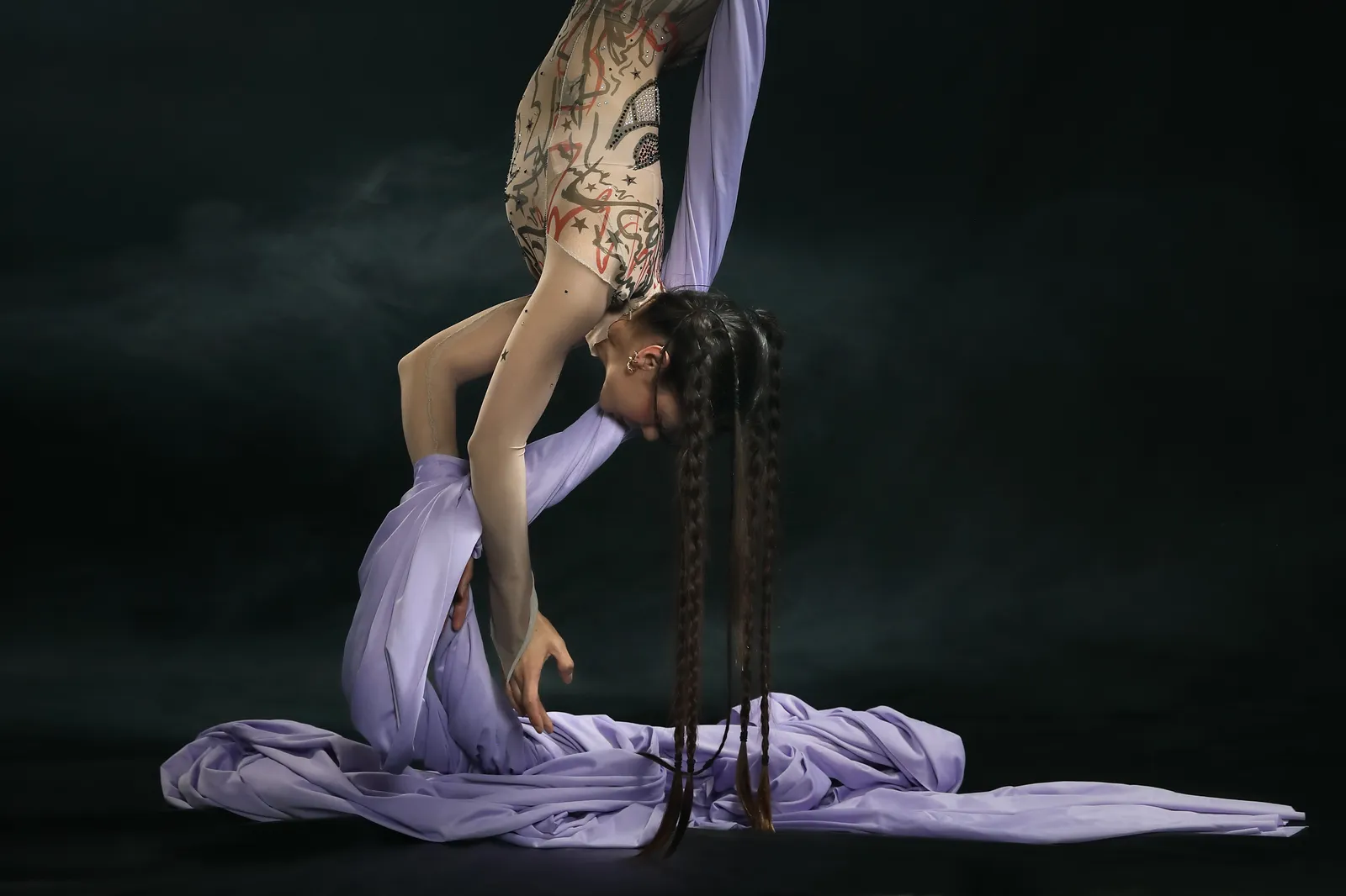 A person performing aerial silks, suspended upside down with long purple fabric draped around them, showcasing intricate tattoos on their arms.