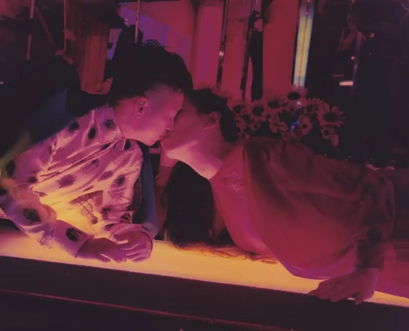A woman and a child leaning toward each other, almost kissing, with colorful lighting casting vibrant hues around them.