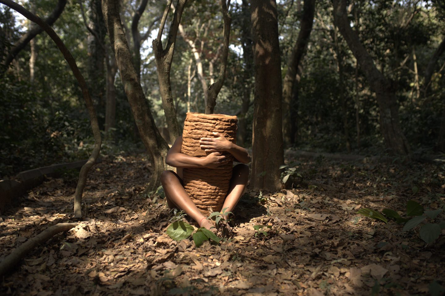 A person sitting on the ground in a forest, holding a large piece of clay or mud in their lap, surrounded by trees and fallen leaves.