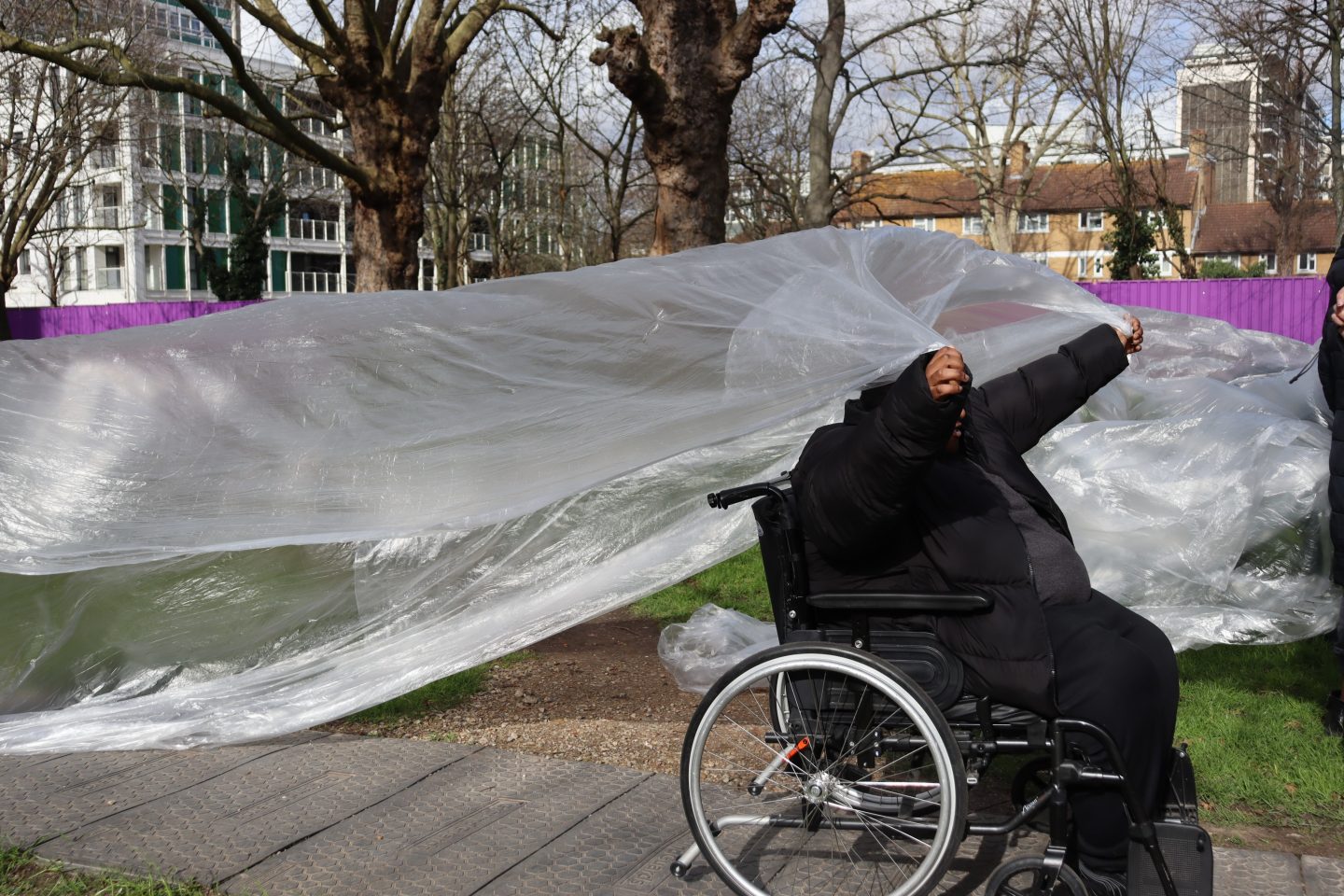 A person in a wheelchair gesturing by a large sheet of plastic draped in an outdoor area with trees and buildings in the background.