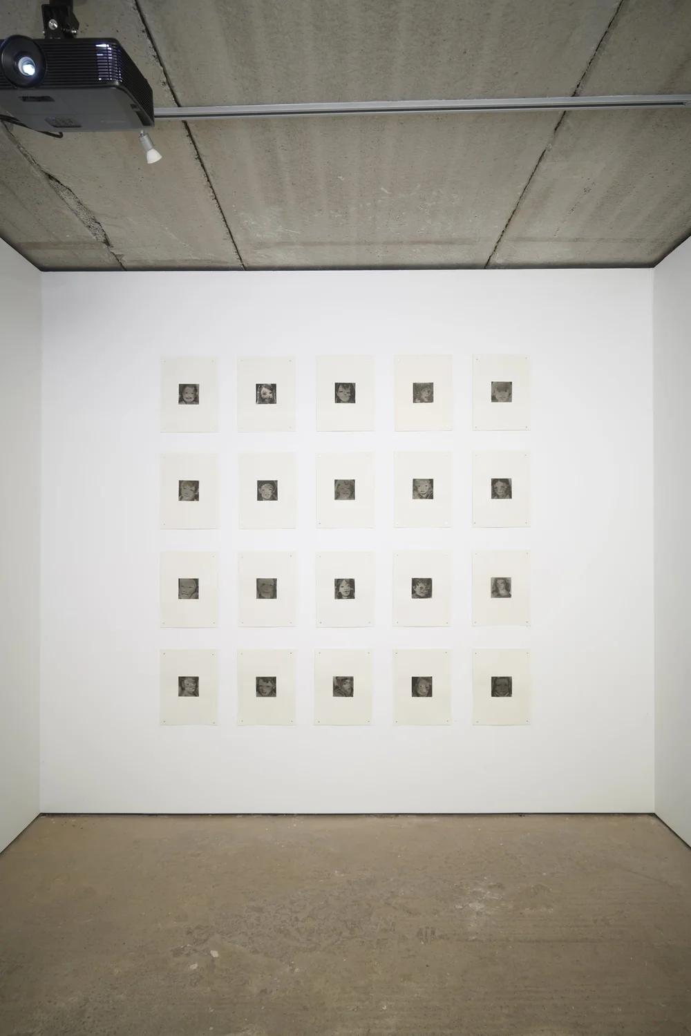 A white wall in a gallery space with a grid of 16 small square artworks displayed. The ceiling has exposed concrete and there is a projector mounted above.