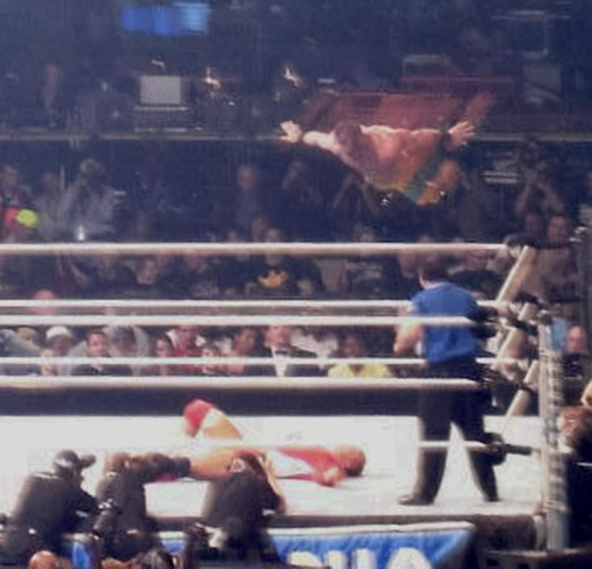 A wrestler diving off the top rope towards another wrestler lying on the mat, with a crowd and referees visible in the background.