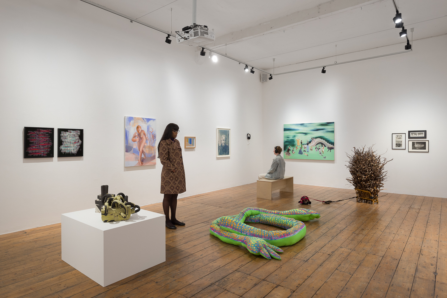 An art gallery interior featuring several artworks on the walls, a woman standing in front of one piece, a man sitting on a pedestal, and an inflatable green serpent-like structure on the floor. | 2026 Nunnery Gallery