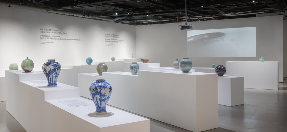A modern art gallery featuring various decorative vases displayed on white pedestals. The walls are plain and there is a projection on the back wall.