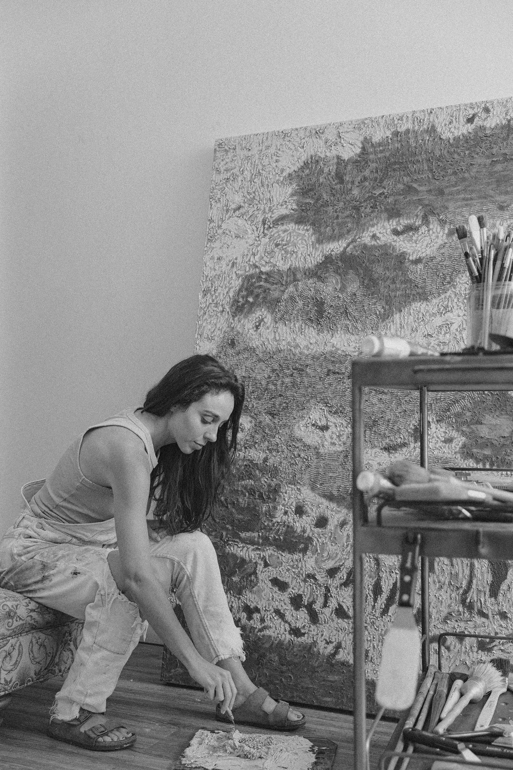 A woman sitting on a sofa, working on her art in a studio filled with painting supplies and an unfinished canvas.