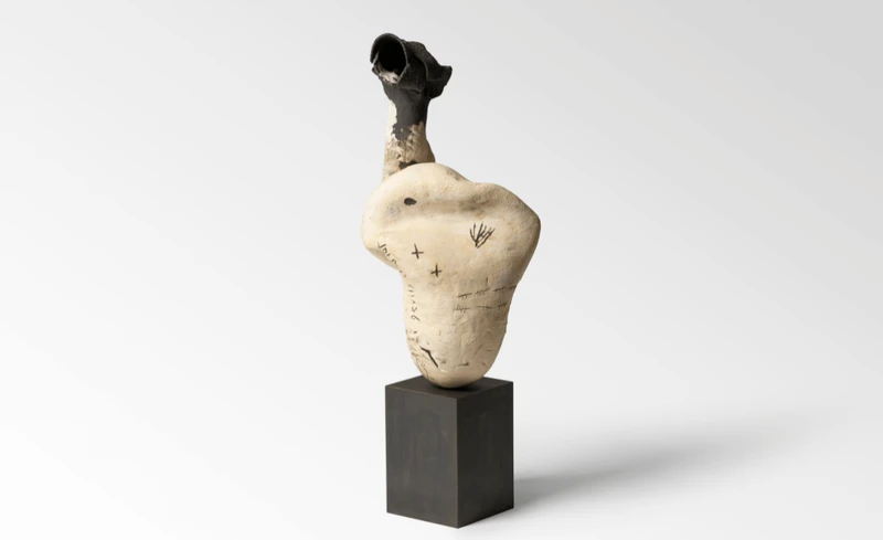 A sculpture resembling an abstract form, featuring a beige-colored body with markings and a black protrusion on top, mounted on a black pedestal.