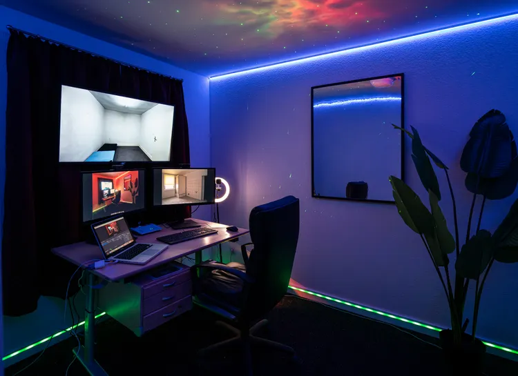 A modern home office setup featuring multiple monitors on a desk, a comfortable chair, and vibrant LED lighting around the room. A large mirror hangs on the wall, and there is a plant in the corner, creating an inviting workspace atmosphere.