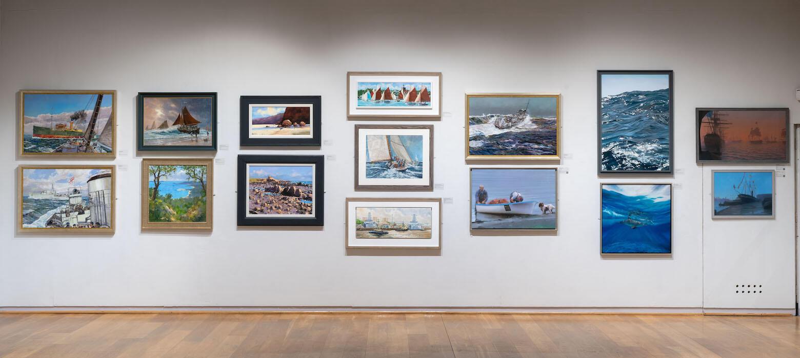 A gallery wall displaying a variety of framed paintings and photographs, mostly depicting marine scenes including boats, waves, and coastal landscapes.