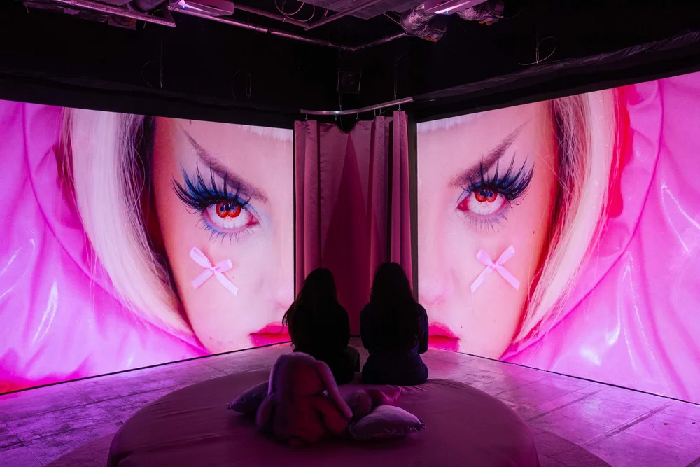 Two individuals sitting on the floor facing large screens displaying an artistic close-up of a face with exaggerated makeup, pastel colors, and prominent eyes.