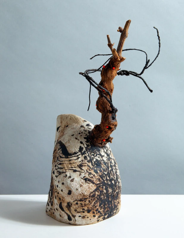 A sculptural piece featuring a textured, abstract shape resembling a head or face, adorned with twisted branches and organic forms protruding from its top. The surface displays a mix of natural colors and patterns.