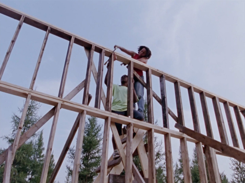 Two individuals are working together on a wooden structure, standing on scaffolding. One person is holding a piece of wood while the other is using a tool to secure it in place. There are trees in the background and the sky is clear.