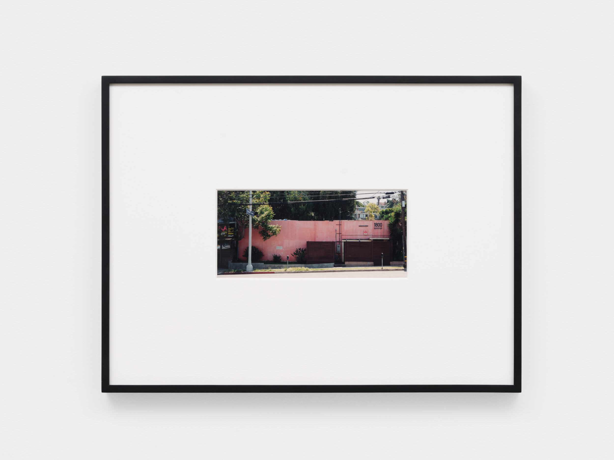 A framed photograph of a pink wall with some greenery and a street in front, displayed on a white background.