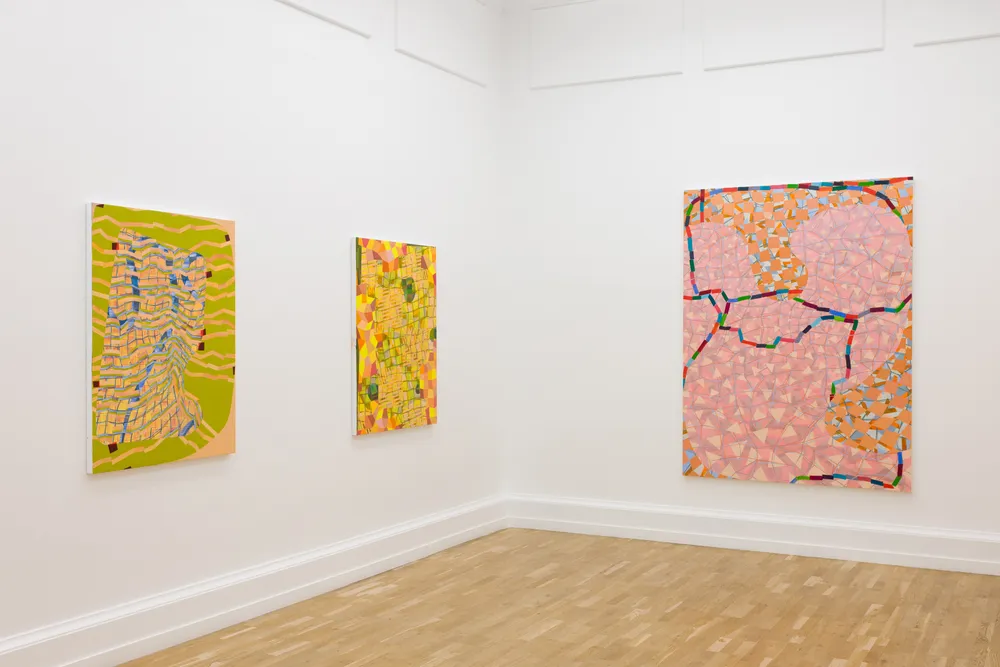 Three abstract paintings are displayed on white walls in a gallery, with one larger painting in the center and two smaller ones on either side. The artworks feature vibrant colors and patterns. The floor is wooden and light-colored, creating an open, minimalist atmosphere.