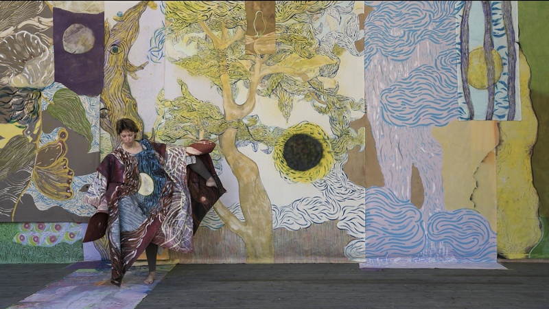 A person wearing a colorful outfit stands in front of a large, abstract mural that features intricate patterns and designs, including trees and waves.