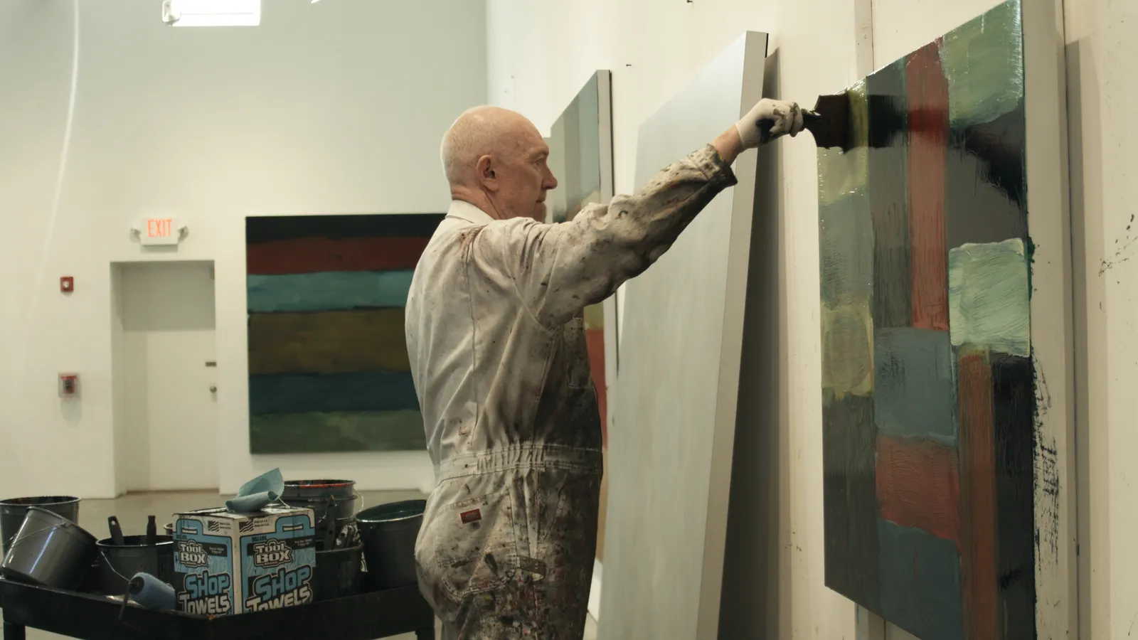 An artist in overalls is applying paint to an abstract canvas on the wall in an art studio, with paint supplies visible nearby.