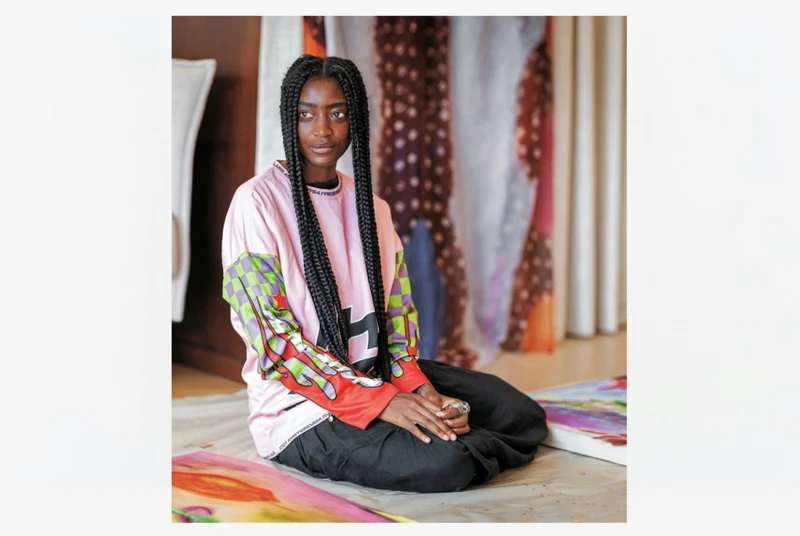 A person with long braided hair sits on the floor, wearing a colorful oversized shirt and black pants. They are surrounded by fabric artworks in a warmly lit indoor setting.