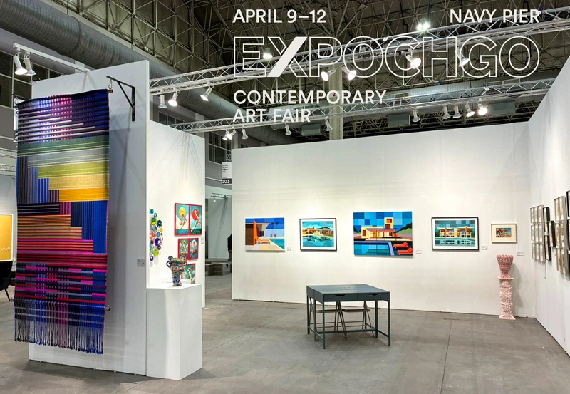An art fair booth featuring colorful artworks on display, including vibrant wall hangings and framed pieces in various styles. A central table is positioned in the gallery space, with additional artwork visible on nearby walls. The event is titled "EXPOCHGO" and takes place from April 9-12 at Navy Pier.