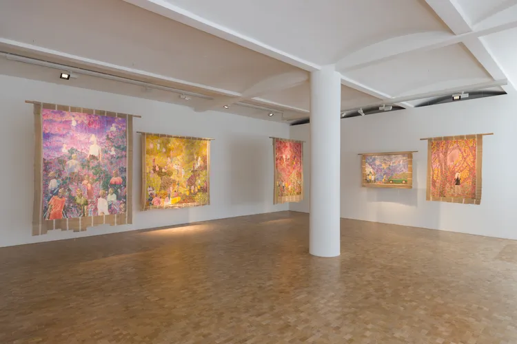 An art gallery with multiple colorful tapestries hanging on the walls, depicting various scenes and figures. The floor is made of light-colored wood, and the space is well-lit with white pillars.