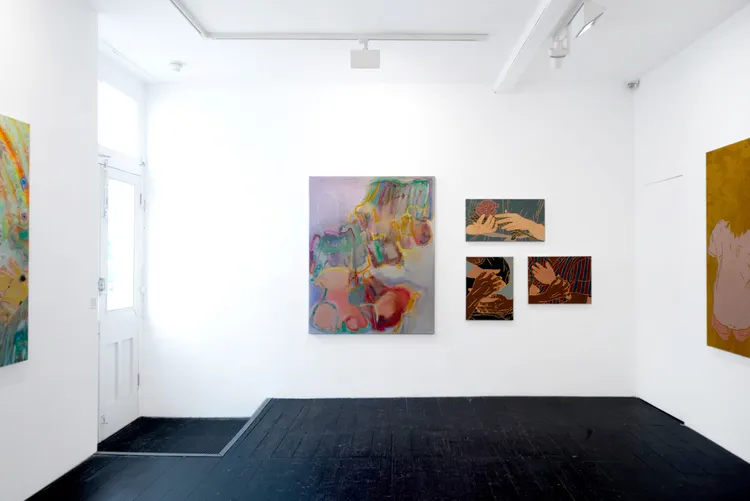 A white-walled art gallery with various paintings displayed. On the left, a large colorful painting is hung, while five smaller artworks are arranged on the right wall. The floor is black and there is a door visible in the background.