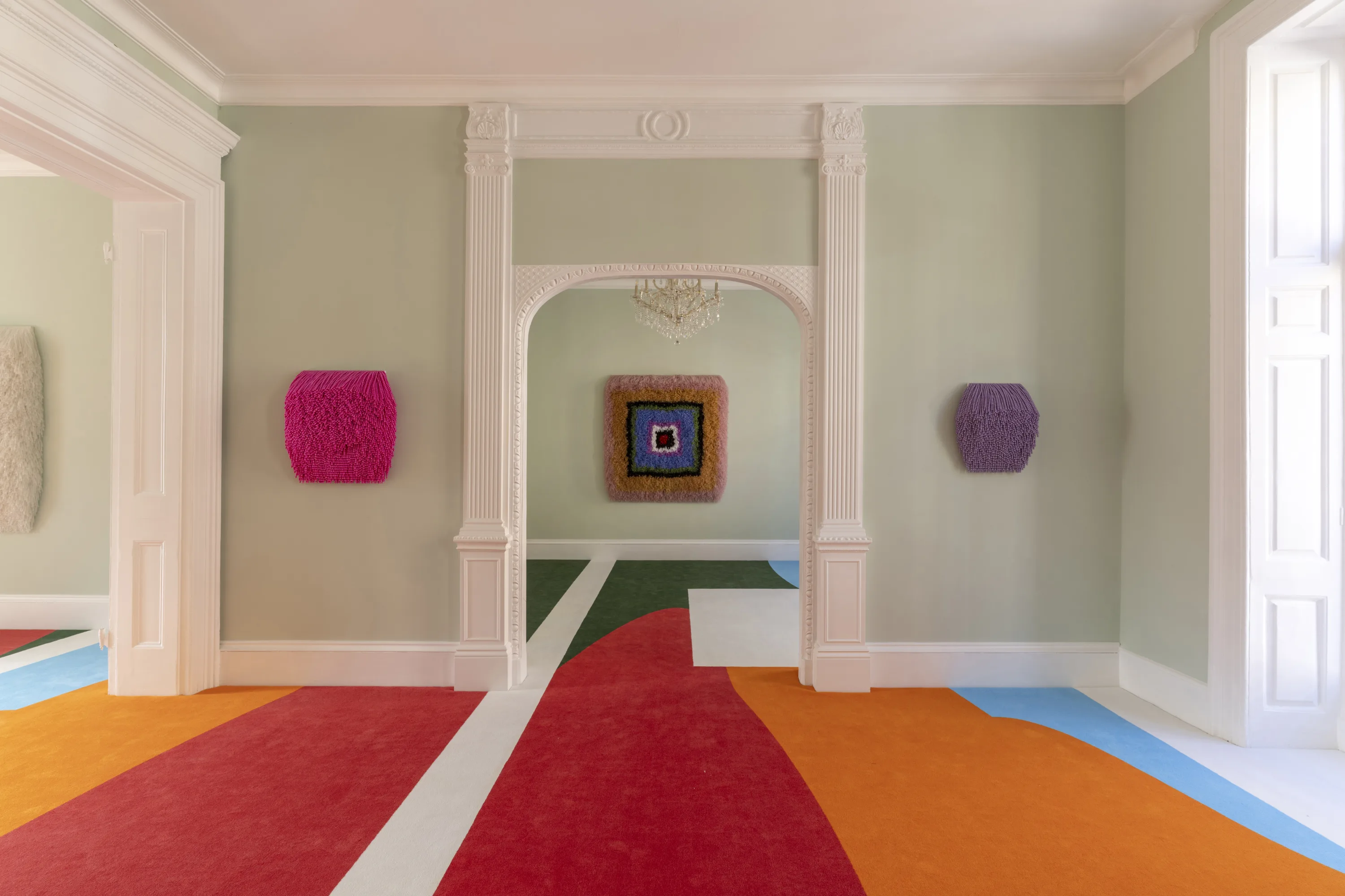 A modern interior space featuring colorful abstract artworks on the walls and a multicolored floor design. The room has tall white columns and a chandelier, with light green walls enhancing the vibrant colors in the artwork and flooring.