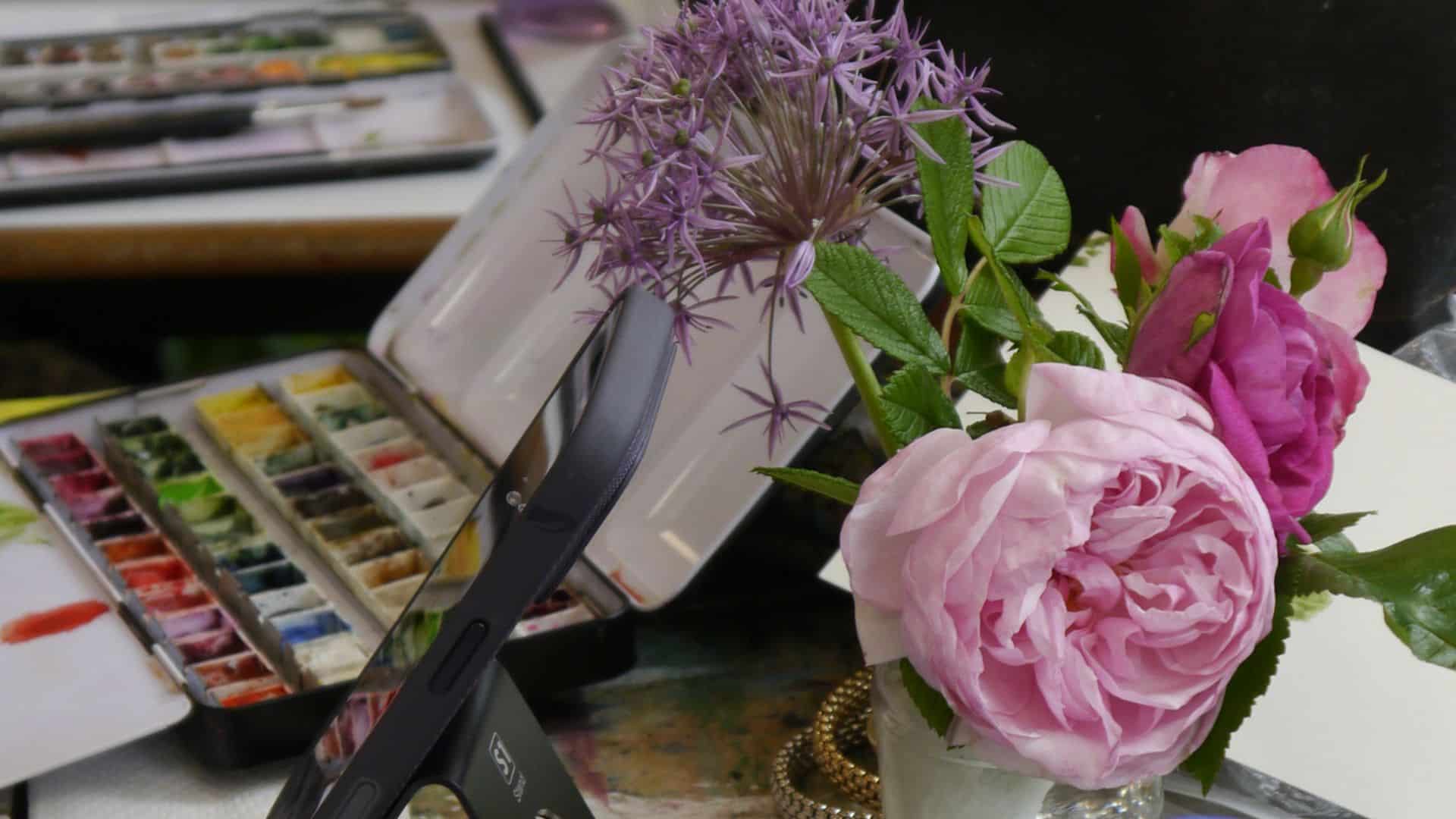 A close-up of a bouquet with pink roses and purple flowers on a table, next to a watercolor paint palette and painting supplies.