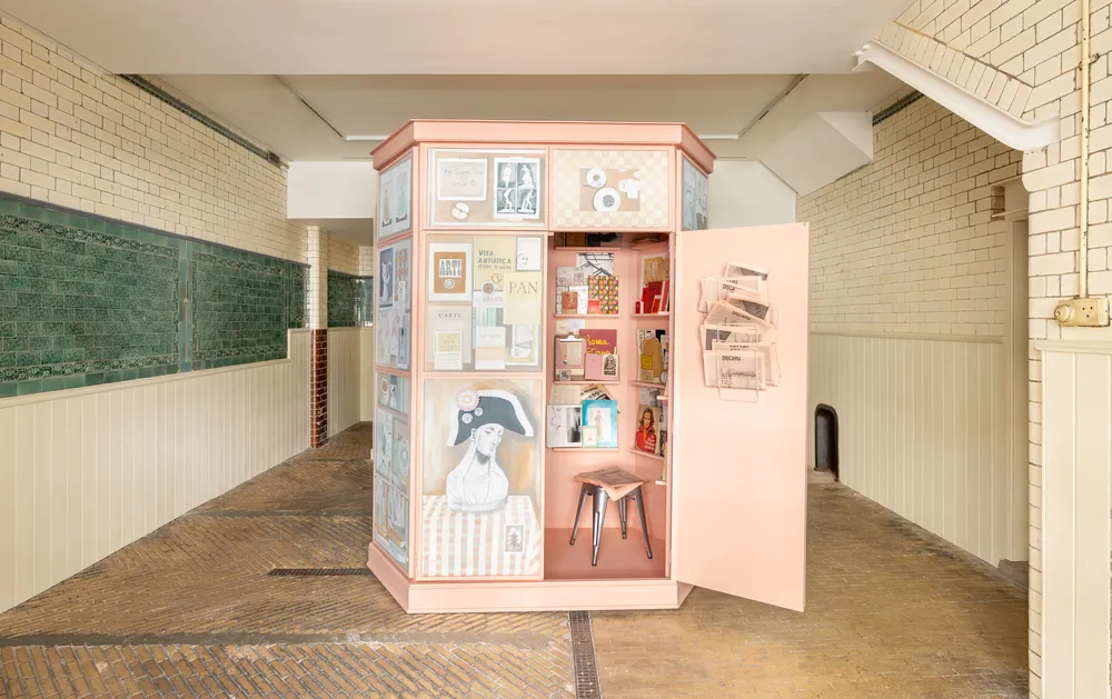 A pink kiosk filled with various items and artwork, located in a corridor with white brick walls and green chalkboards. The kiosk door is open, revealing shelves lined with books, decorative objects, and art pieces.