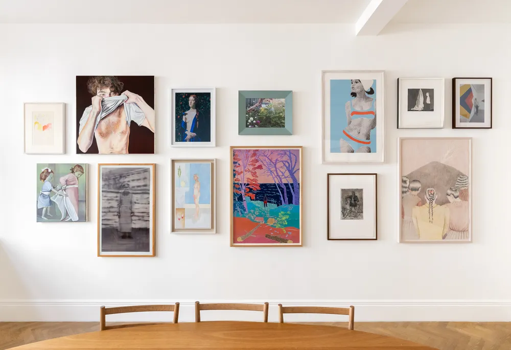 A gallery wall featuring various framed artworks, displayed above a wooden table. The pieces include a mix of styles and subjects, such as abstract designs and figurative representations.
