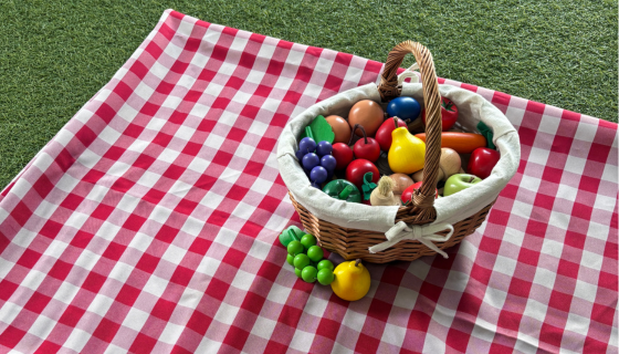 A woven basket filled with colorful wooden fruits and vegetables sitting on a red and white checkered picnic blanket.