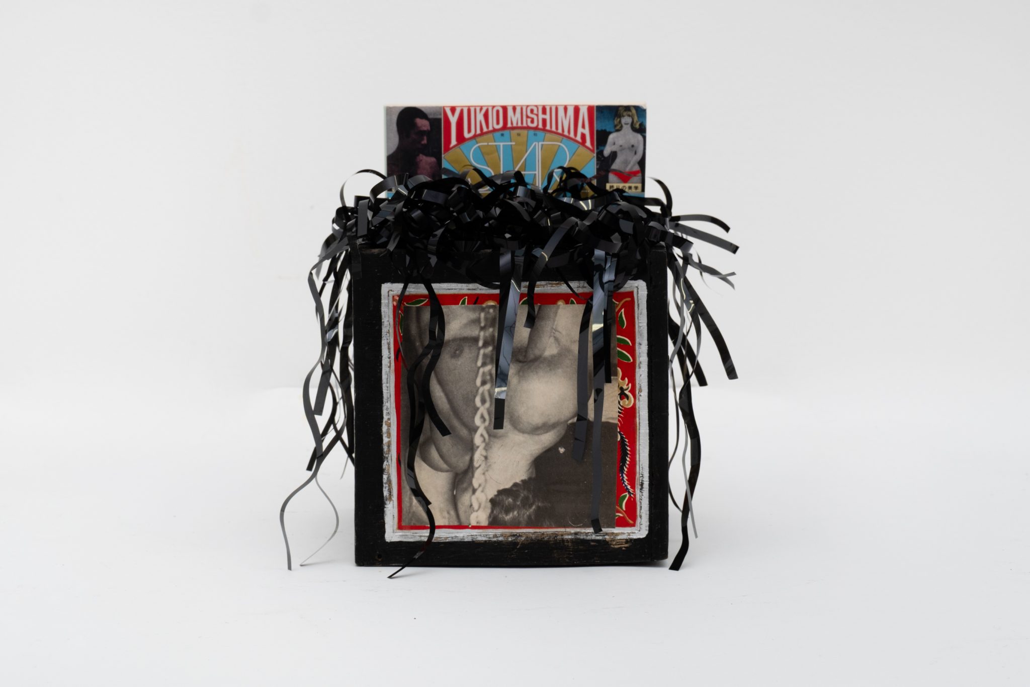 A decorative box with black and red fringe, containing a black and white photo of a person along with a magazine cover featuring the name "Yukio Mishima."