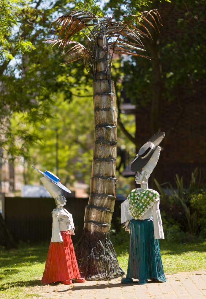Two decorative figures made of metal, one dressed in a red skirt and blue hat, the other in a green and white checkered top with a black hat, stand beside a tall palm tree sculpture in a park.