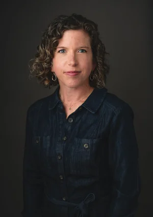 A woman with curly hair wearing a denim shirt, standing against a dark background.