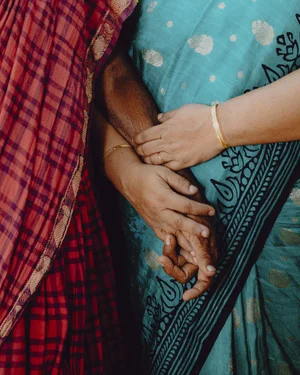 Two women wearing traditional attire, standing close together, with their arms intertwined. One woman has a red and black checked garment, while the other wears a blue sari adorned with intricate patterns. Their hands are gracefully clasped together. | 2026 Photofusion