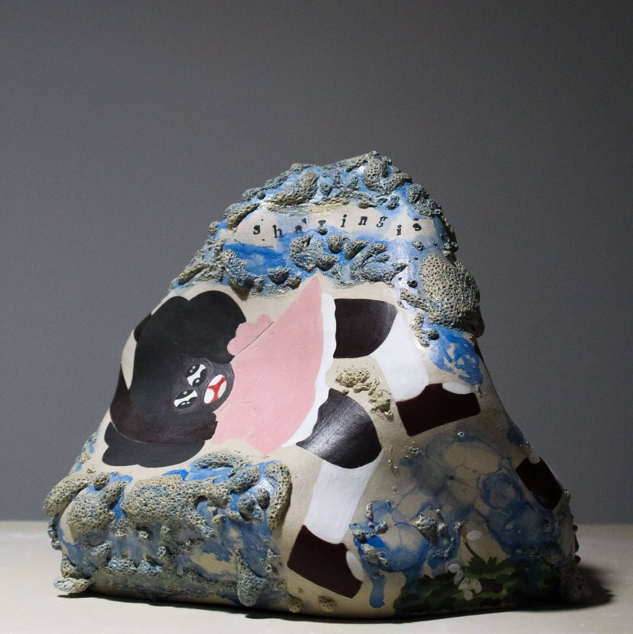 A sculptural piece resembling a rock, featuring various textures, colors, and abstract designs including a painted figure with black hair and red lips.