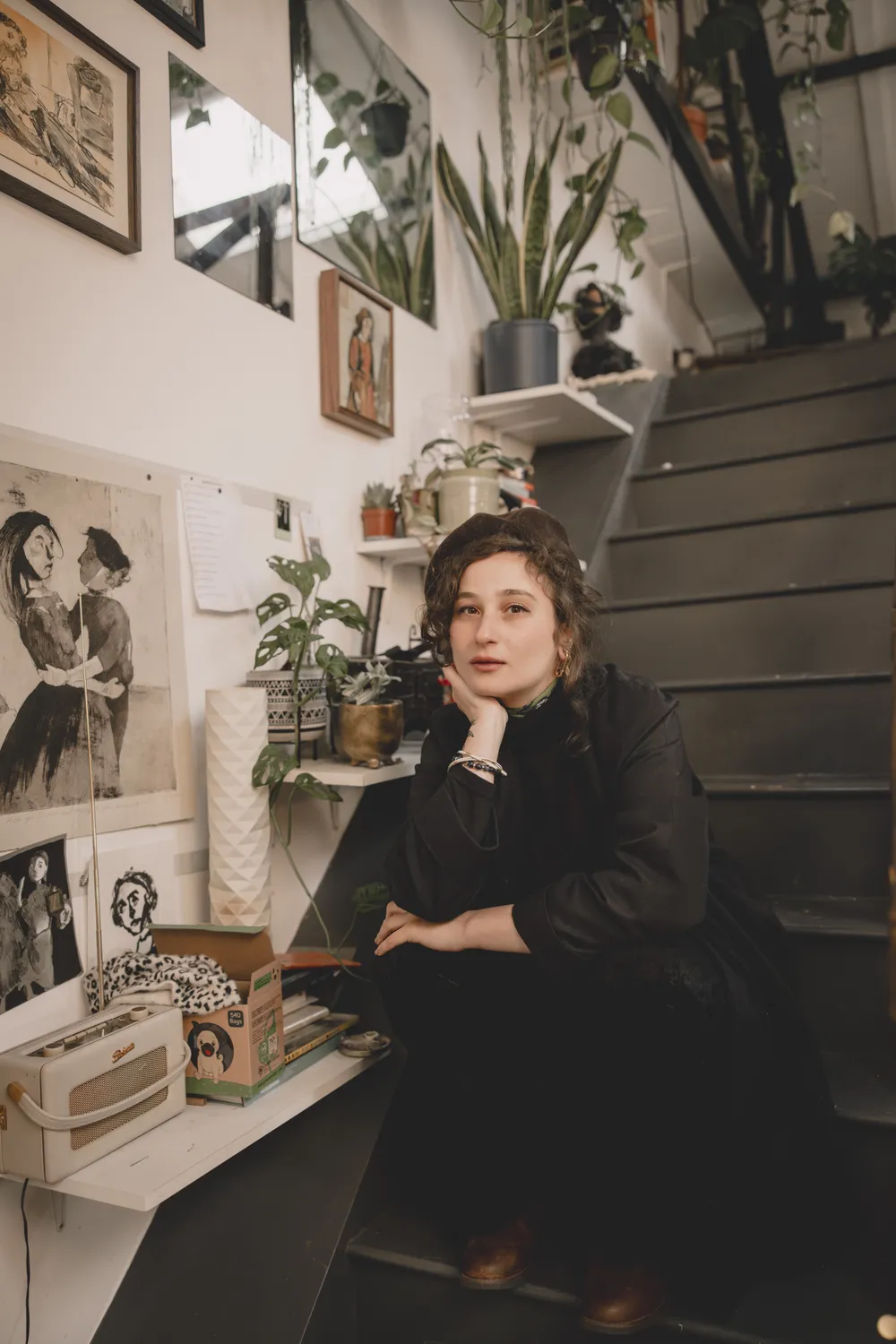 A person sitting on stairs in a stylish indoor setting, surrounded by plants and artwork on the walls.