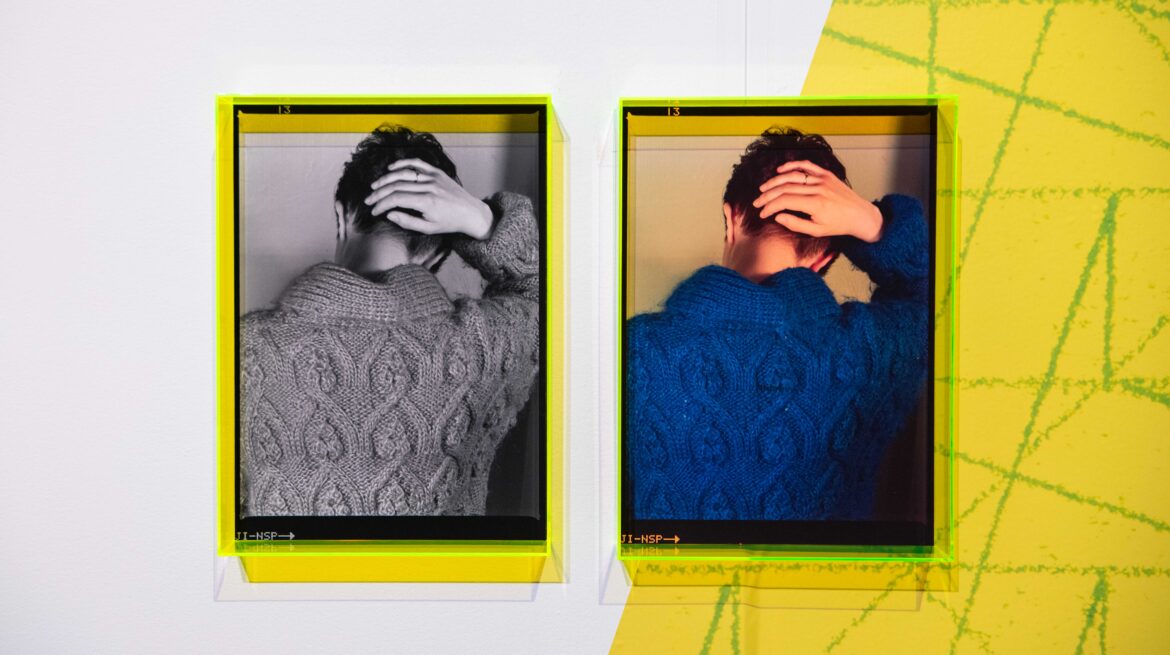Two framed images of the same person from behind, one in black and white and the other in blue, both featuring a textured sweater and a hand on the head. The frames are yellow and show a geometric pattern on the wall.