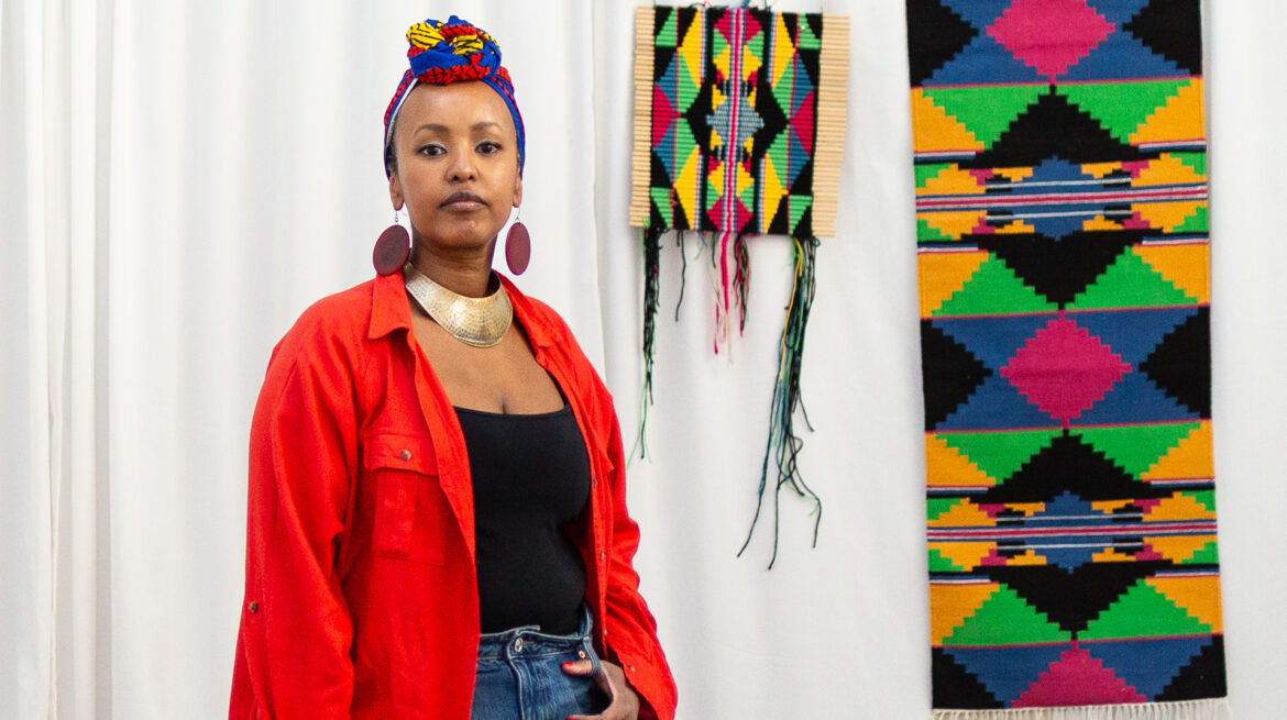 A stylish woman stands in front of colorful woven wall hangings, wearing a bright red jacket and large earrings.