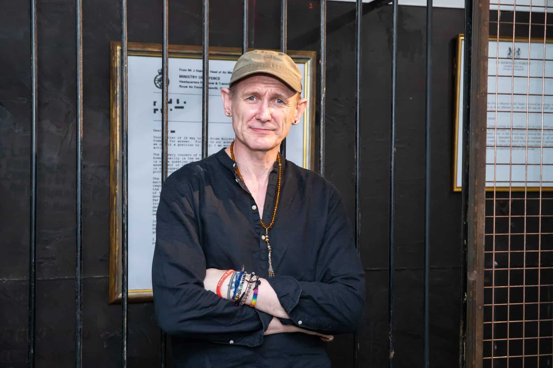 A man wearing a cap and black shirt stands with folded arms behind black metal bars, with framed documents on the wall behind him. | 2026 Royal Air Force Museum London