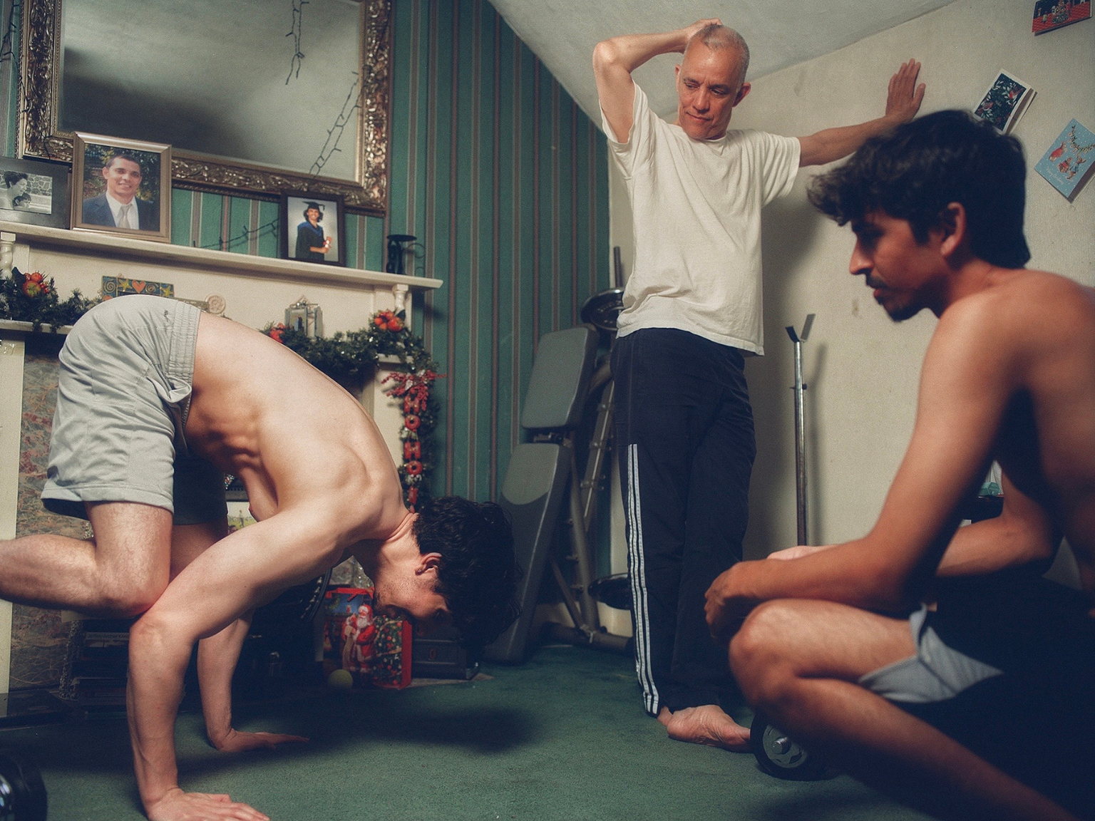 Two men observe a third man performing an exercise on the floor. The room is decorated with family photos and has exercise equipment in the background.