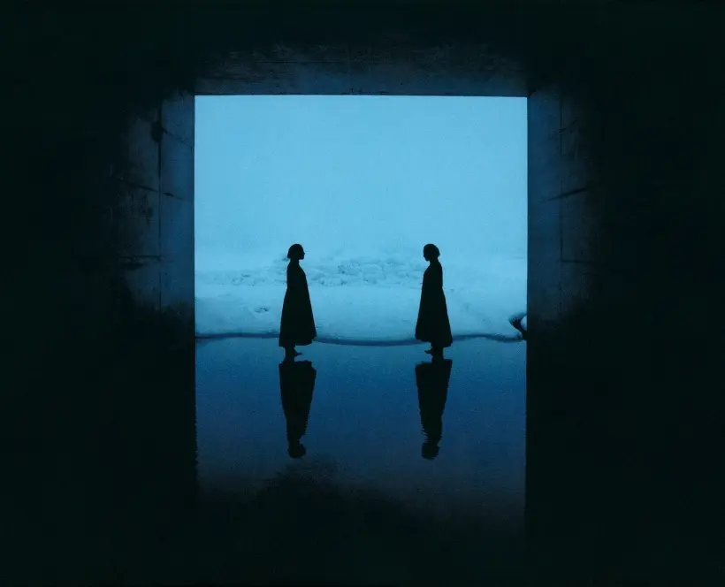 Two figures in dark silhouettes stand facing each other in a dimly lit space, with a blue background and reflections in water.