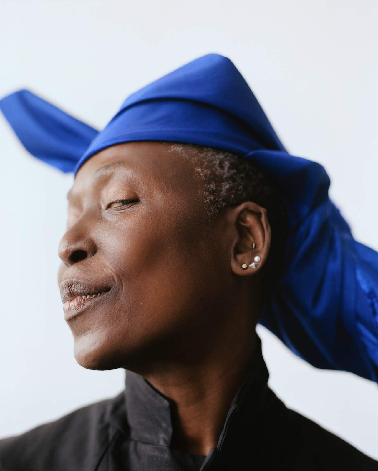 A person with short hair wearing a striking blue hat and a black top, looking slightly to the side.